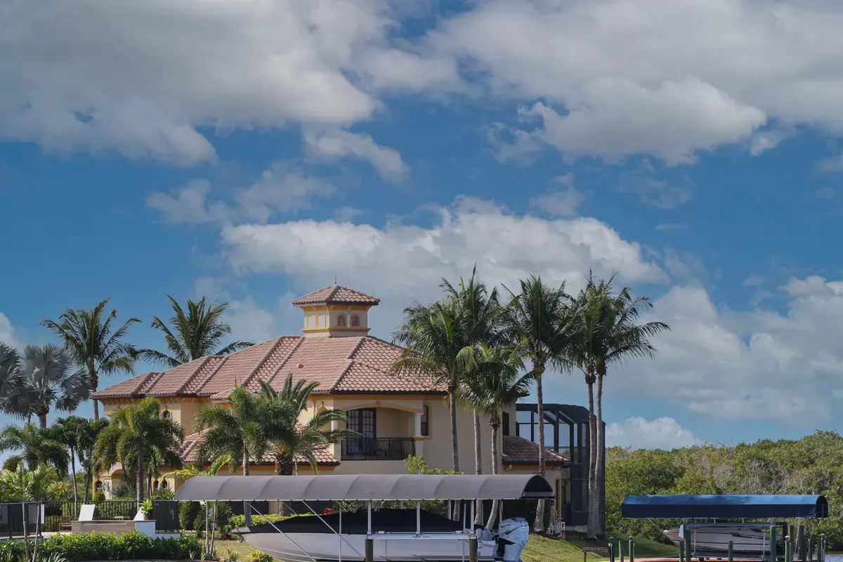 Tile-roofed waterfront home with palm trees and boat lifts under a bright blue sky